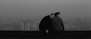 En mand lægger hovedet på ryggen af en anden mand, begge sidder højt over byen på et tag.