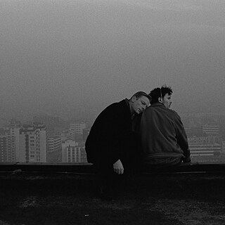 En mand lægger hovedet på ryggen af en anden mand, begge sidder højt over byen på et tag.