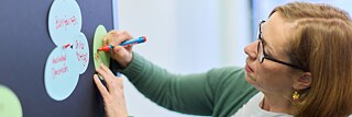 Eine Frau mit schulterlangen Haaren und Brille schreibt etwas auf einen bunten Zettel, den sie an eine Pinnwand heftet. 
