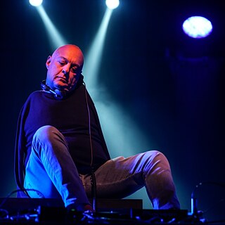 Un DJ sin brazos toca música con los pies. El DJ está sentado en un escenario, iluminado por dos focos en el fondo. Lleva auriculares y viste ropa oscura.