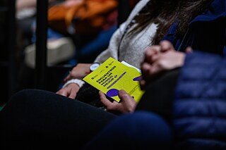 Una persona sostiene un folleto amarillo con el título „Encuentros para un mundo mejor“ en las manos. La escena parece ser en un auditorio o evento, ya que se pueden ver más personas desenfocadas en el fondo.