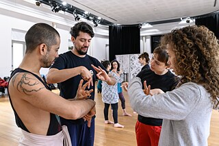 La imagen muestra a un grupo de personas en un estudio de danza o movimiento. Cuatro personas están en primer plano y parecen estar concentradas en gestos de legua de señas con las manos, posiblemente como parte de un ejercicio o taller. En el fondo se pueden ver más personas que también participan en la actividad. La sala es luminosa y espaciosa con suelo de madera y grandes ventanas.