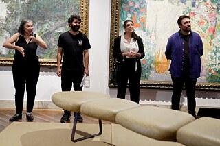 Cuatro personas están de pie frente a dos grandes pinturas en un museo. La persona más a la izquierda está usando el lenguaje de señas. Las otras tres personas están paradas una al lado de la otra y parecen estar escuchando o hablando. En primer plano hay un banco beige.