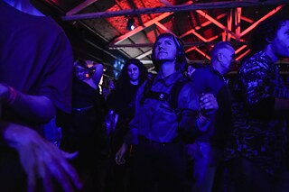 Un grupo de personas baila en una habitación oscura con iluminación azul y roja. Algunas personas llevan auriculares. El techo tiene vigas de madera visibles que también están iluminadas.