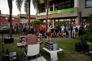 Un evento al aire libre frente al Goethe-Institut Uruguay. Un grupo de personas está de pie y sentadas frente al edificio mientras un músico toca un teclado. En el fondo se puede ver un cartel verde con la inscripción „Goethe-Institut Sprache, Kultur, Deutschland“. La escena tiene lugar en una zona ajardinada rodeada de palmeras y cadenas de luces.