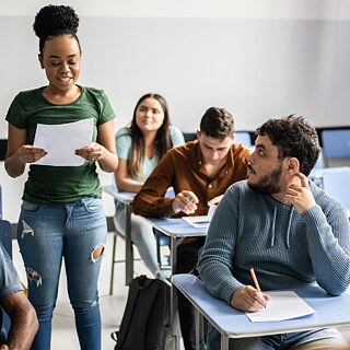 Eine Schülerin liest den Klassenkameraden etwas vor, diese machen sich Notizen. Im Vordergrund steht unscharf die Lehrerin.