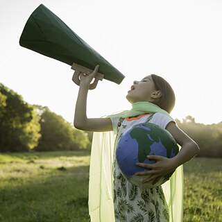 Eine junge Öko-Aktivistin steht auf einer idyllischen Wiese. Sie hält einen Globus im Arm und spricht in ein Megafon.