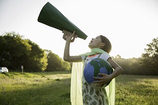 Eine junge Öko-Aktivistin steht auf einer idyllischen Wiese. Sie hält einen Globus im Arm und spricht in ein Megafon.