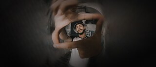 Person mit Fischerhut wird von zwei Händen in Rechteckform eingerahmt, steht in einem schwach beleuchteten Raum.