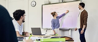 Žena stojí u interaktivní tabule a něco vysvětluje.