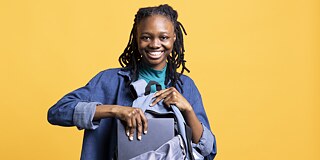 Eine lächelnde junge Frau mit Dreadlocks steht vor einem gelben Hintergrund und steckt einen Laptop in ihren grauen Rucksack.