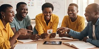 Five students are sitting at a table and working on various documents.  They are cheerful and laughing.