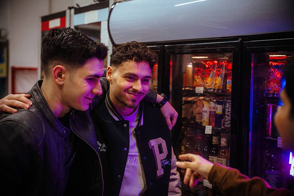 Im Kiosk: Yasin (Mert Dincer) und Dominik (Angelo Alabiso) haben sich gegenseitig ihre Arme um die Schultern gelegt und sehen Berkan (Dennis Kharazmi) grinsend an.