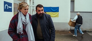 Karla (Franziska Machens) und Eren (Sahin Eryilmaz) stehen vor der Flüchtlingsunterkunft, während im Hintergrund eine Person einen Karton trägt.