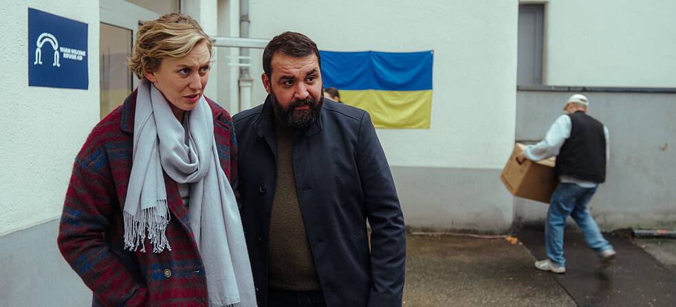 Karla (Franziska Machens) und Eren (Sahin Eryilmaz) stehen vor der Flüchtlingsunterkunft, während im Hintergrund eine Person einen Karton trägt.