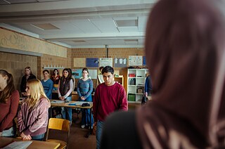 Ahmad (Rasmi Mohammed Nasrallah) verlässt wütend den Klassenraum, während seine Mitschülerinnen und Mitschüler mit gefalteten Händen an Ihren Plätzen stehen.