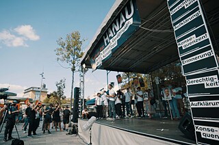 Reporter und Fernsehteams am stehen am Jahrestag vor der Bühne der Gedenkveranstaltung in Hanau. Auf der Bühne stehen mehrere Personen, eine Person spricht.