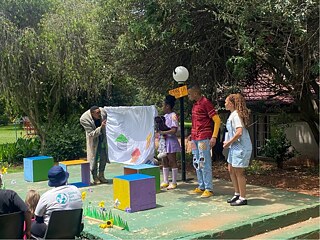 Children from Amandla Street perform on a small outdoor stage decorated with colorful blocks and flowers, holding a hand-drawn sheet, while an audience watches from chairs surrounded by greenery.