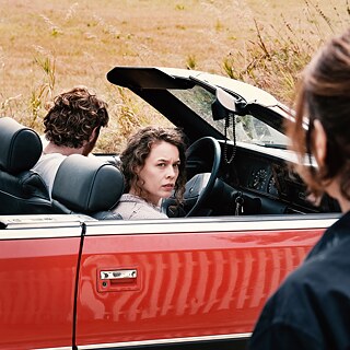 Filmstill: Two people, perhaps a couple, in a red cabriolet are passing a woman
