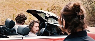 Filmstill: Two people, perhaps a couple, in a red cabriolet are passing a woman