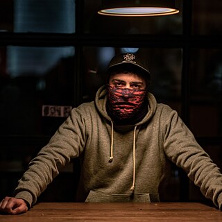 Person mit Kapuze, Mütze und Gesichtsbedeckung steht hinter einem Holztisch in einem schwach beleuchteten Raum; warmes Licht fällt von oben.