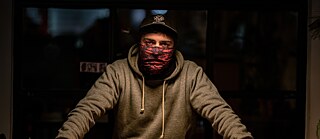 Person mit Kapuze, Mütze und Gesichtsbedeckung steht hinter einem Holztisch in einem schwach beleuchteten Raum; warmes Licht fällt von oben.