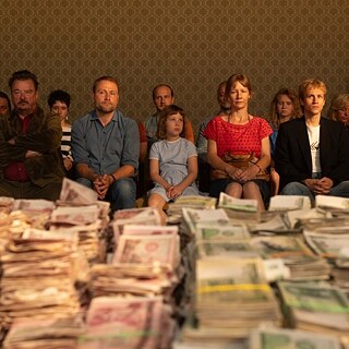 Dozens of people are seated in a room. On a table in front of them lies a lot of money