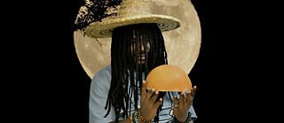 SOUROUNDOU – EINTOPF | In einer anderen Komposition wird der Mann von einem Vollmond eingerahmt, einer Metapher für Hoffnung und Wiedergeburt. Er trägt einen Strohhut aus der Sahelzone und hält eine goldfarbene Kalebasse in der Hand, deren Farbton an die goldene Farbe der deutschen Flagge erinnert und den interkulturellen Dialog zwischen Afrika und Europa symbolisiert. Sein teilweise beleuchteter Körper hebt sich  von einem tiefschwarzen Hintergrund ab und  unterscheidet so den unsichtbaren kreativen  Prozess von der sichtbaren Darbietung,  die mit dem Publikum geteilt wird. In der Mitte positioniert, wird der Mann  zur Allegorie aller afrikanischen  Künstler, die zwar unterschiedliche kreative  Identitäten haben, aber durch eine gemeinsame Vision vereint sind.  Der Hut und die Zweige, die mit dem  Mond verschmelzen, schaffen eine symbolische Verbindung von Natur,  Spiritualität und Fantasie und verstärken  die surreale und kontemplative  Atmosphäre des Werks. Sein Blick, der in die Zukunft gerichtet ist,  verkörpert die Hoffnung einer neuen Generation  von Künstlern, die durch das  Souroundou-Projekt zusammengebracht wurden und die  Begegnung der Kulturen, die Erinnerung an  Traditionen und das Licht der künstlerischen Erneuerung Afrikas  feiern. 