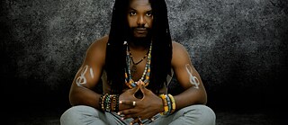 SOUROUNDOU – EINTOPF | The image depicts a man with dreadlocks,  a living embodiment of African identity,  seated cross-legged against a dark,  textured background. His serene yet  powerful presence evokes a balance  between tradition and modernity. He wears multiple necklaces and  bracelets, adorned with colorful beads and  motifs inspired by the Sahel and West  Africa, expressing a deep cultural  rootedness. His joined hands, positioned  before him in a gesture of creative meditation, reveal a moment of inner focus and spiritual  connection with his art. White symbols painted on his shoulders represent cultural fusion and the harmonious  dialogue between different peoples. At his feet lie two symbolic objects: a microphone,  embodying modern music, and a calabash, a traditional instrument and emblem of the  Souroundou project. Together, they bridge ancestral oral heritage and contemporary artistic  expression. The man’s gaze, fixed on  these objects, conveys the meeting point  between heritage and creation.