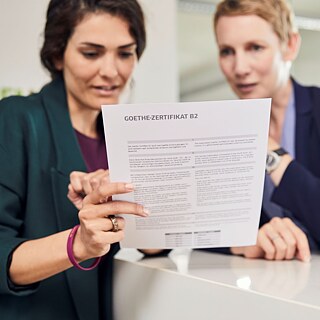 Gabriela hat die Prüfung bestanden. Sie hält das Goethe-Zertifikat B2 in der Hand und sichtet mit der Lehrkraft die Ergebnisse.