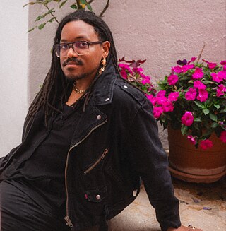 A foto mostra Alex d'Alva Teixeira sentado no chão, apoiando-se com uma mão e olhando para a câmara. Ao fundo, há plantas cor-de-rosa em vasos e hera a crescer na parede.