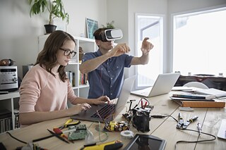 Ein Vater sitzt mit seiner Tochter zu Hause am Tisch, er trägt eine VR-Brille und zeigt mit den Händen in die Luft, sie programmiert mit einem Laptop den Virtual-Reality Simulator.