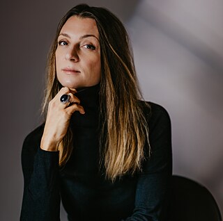 A foto mostra a artista Julia Lehmann, com a cabeça ligeiramente apoiada nas mãos, usando um anel com uma pedra preta no dedo e olhando para a câmara.