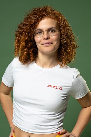 A foto mostra Maria Caetano Vilalobos com as mãos na cintura, olhando para a câmara diante de uma parede verde.