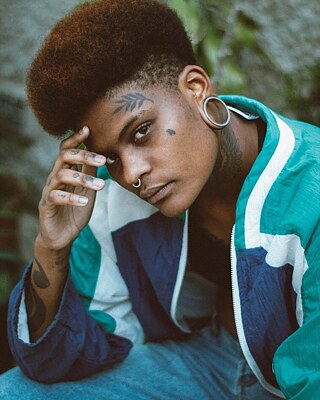 A imagem mostra Naina Costa com uma mão no rosto, olhando para a câmara. Ao fundo, vêem-se plantas.