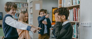 Drei Kinder zeigen mit dem Finger auf ein anderes Kind, das sich die Hände vors Gesicht hält.