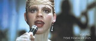 Person in elegantem, goldfarbenem Outfit hält ein Mikrofon auf einer Bühne, im Hintergrund unscharfe Musiker und Instrumente, darunter eine Gitarre. 