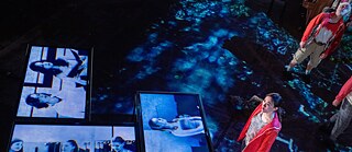 The 5th Season – Fat Tuesday:  Eine Performerin in einer roten Jacke blickt nach oben auf mehrere große Videobildschirme auf dem Boden, die schwarz-weiße Gesichter von Menschen im Gespräch zeigen. Die Beleuchtung ist kühl und blau getönt.