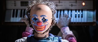 The 5th Season – Fat Tuesday:  A person wearing a brightly lit clown mask with glowing blue outlines plays an electric keyboard. The scene is dimly lit, with a microphone visible nearby.