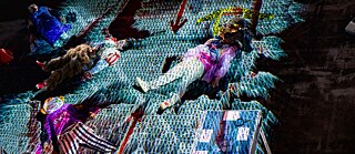 The 5th Season – Fat Tuesday:  Several performers lie on a stage covered in a patterned light projection. They wear colorful costumes and masks, with props like hats and a table of objects scattered around.