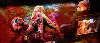 The 5th Season – Fat Tuesday:  A performer with curly blond hair sings passionately into a microphone under colorful stage lights, while another person plays the piano beside them. A third performer crouches nearby in dramatic lighting.