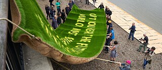Large green sign made from grass being lowered down onto the shore of the river Thames, with photographers taking photos of it