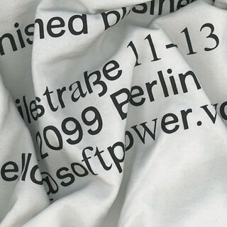 Weiße, zerknitterte Stofffläche mit schwarzem Text: „softpower“, „2099 Berlin“, „11–13“
