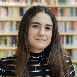 Porträt von Maria Franzky, einer Mitarbeiterin mit langen dunklen Haaren, vor einem Hintergrund mit Bücherregalen. Sie trägt ein schwarzes Oberteil mit gelben und weißen Streifen sowie eine Halskette mit kleinem Anhänger.