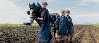 Drei Personen in Arbeitskleidung stehen auf einem Acker neben einem gescheckten Pferd; im Hintergrund weite Landschaft. 