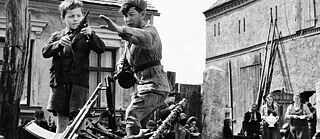 Black and white image that shows a shoulder trying to take a gun from a boy. There is a farm house and farming people in the background.