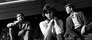Black and white image of a young white woman sitting between two white men sitting on the ground outside.
