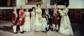 Colour image of children and adults in historical royal attire standing in one row.