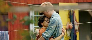 Zwei Menschen umarmen sich innig vor einem roten Holzhaus.