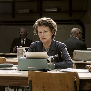 Colour image of an older white woman behind a table watching something outside the picture frame.